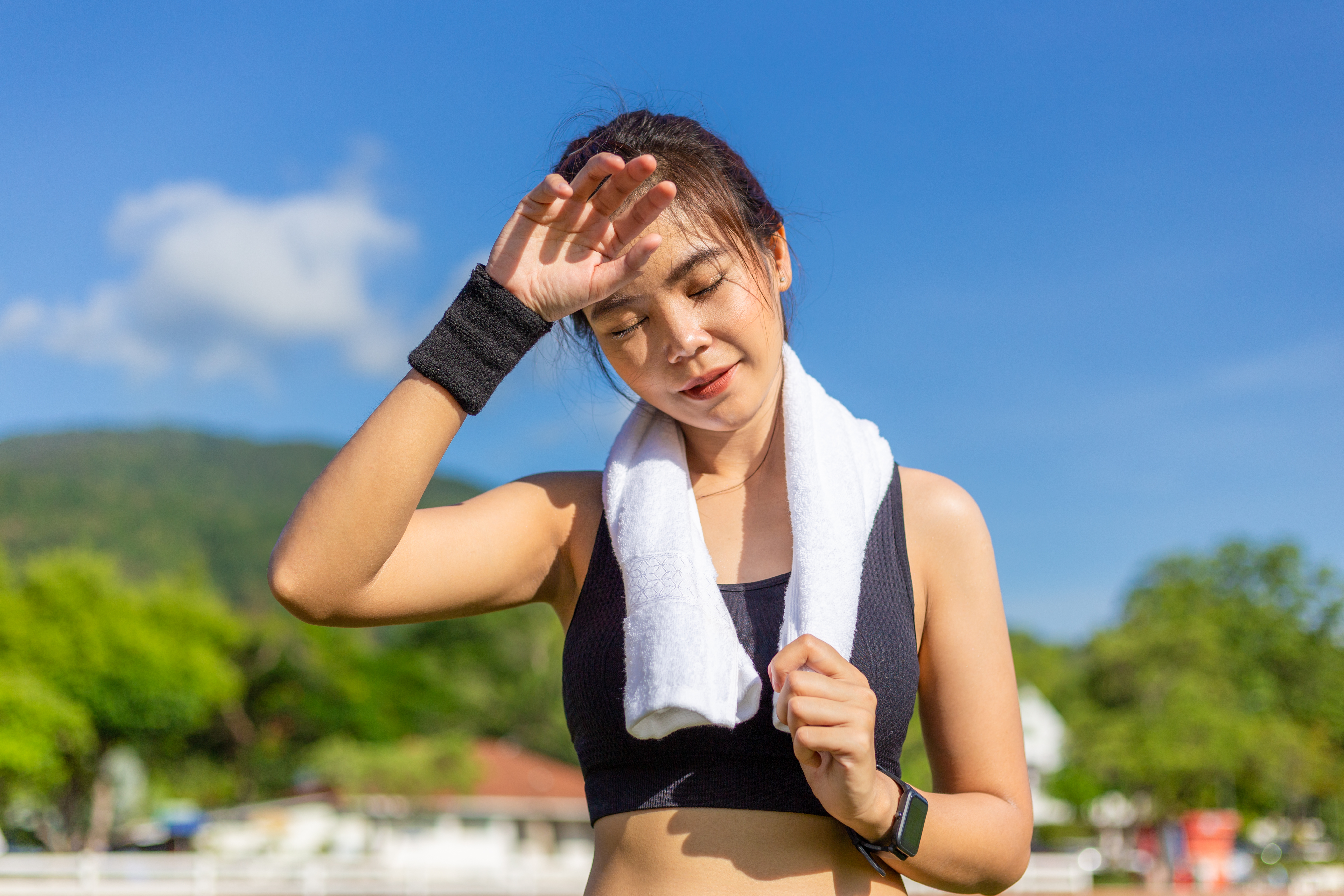 Beautiful young Asian woman stops to wipe off her sweat after her morning exercise run at an outdoor running track on a bright sunny day